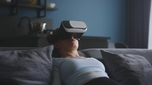 Woman in virtual reality googles enters metaverse immersive expirience via headset interface resting on sofa in minimalistic interior. Girl gaming in cyber space. Futuristic concept.