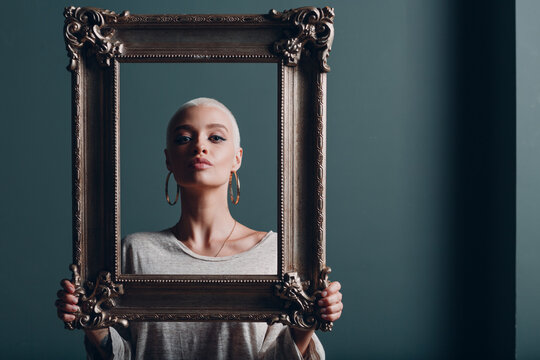 Millenial young woman with short blonde hair holds gilded picture frame in hands behind her face portrait