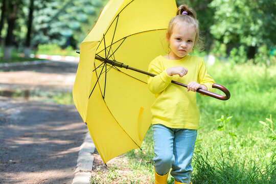 a little cheerful girl walks after the rain in rubber boots and under a yellow umbrella in a warm autumn in the park