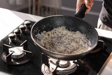 Close up of professional Chef cook hands roasts champignons mushrooms with cream in wok pan for Mediterranean cuisine. 