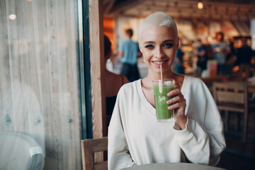 Portrait of young smiling millenial european short haired woman with green smoothie at cafe. Beautiful happy blonde girl indoor.