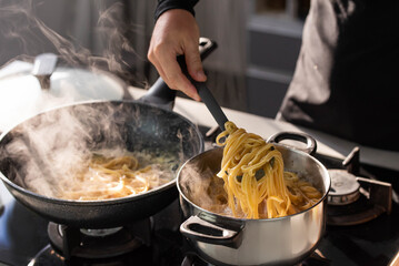 Professional chef cook making Italian Tagliatelle pasta with mushrooms and cream at modern kitchen gas stove in wok pan and pot.