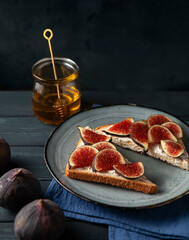 Two toasts on the plate with cream cheese and figs and jar of honey on the dark blue wooden table, selective focus