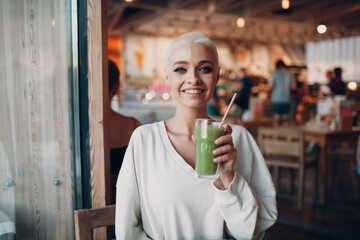 Portrait of young smiling millenial european short haired woman with green smoothie at cafe. Beautiful happy blonde girl indoor.