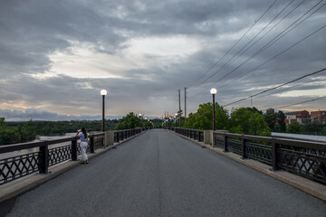 View down bridge roadway deck at dusk with lone woman looking at the river below, lit street lamps on bridge railing, dramatic cloudy sky