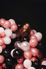 A girl with long hair in a black shiny dress celebrates her birthday with a lot of colorful balloons isolated on a black background. Copy space.
