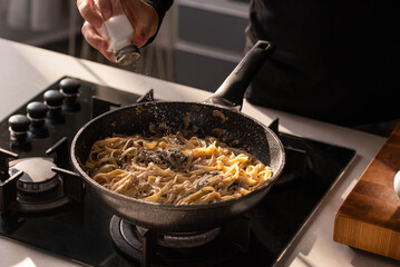 Professional chef cook making vegetarian Italian Tagliatelle pasta with mushrooms and cream at modern kitchen gas stove in wok pan.
