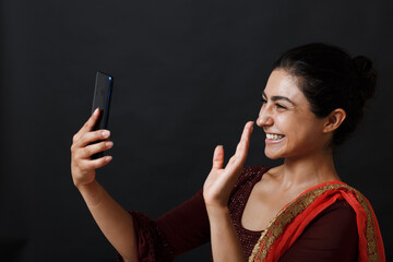 Happy indian woman having video call on mobile phone. Brunette in traditional sari clothes of red and gold colour. Beautiful smiling dark haired young woman. Isolated on dark background. Studio shoot