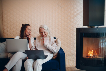 Senior and young woman using computer laptop at sofa. Mom and daughter sitting by fireplace indoor.