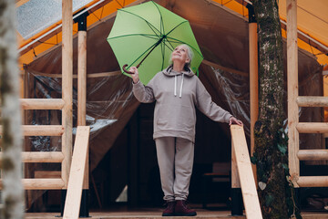 Elderly senior woman with umbrella at glamping camping tent. Modern vacation lifestyle concept