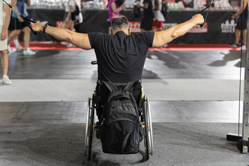 Fototapeta na wymiar Man Sitting in a Wheelchair with a Backpack attached doing Arm and Shoulder exercises with Gym Equipment