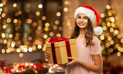 Obraz na płótnie Canvas christmas, holidays and people concept - happy smiling teenage girl in santa helper hat holding gift box over festive lights at home background