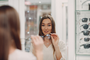 Close up of gorgeous young woman smiling picking and choosing glasses at the optician corner at the shopping mall. Happy beautiful woman buying eyewear eyeglasses at the optometrist.