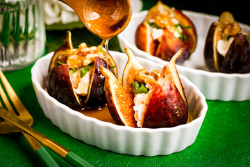Baked figs with cheese and nuts in white dishes on a green table. Nearby are a fork and a knife. High quality photo
