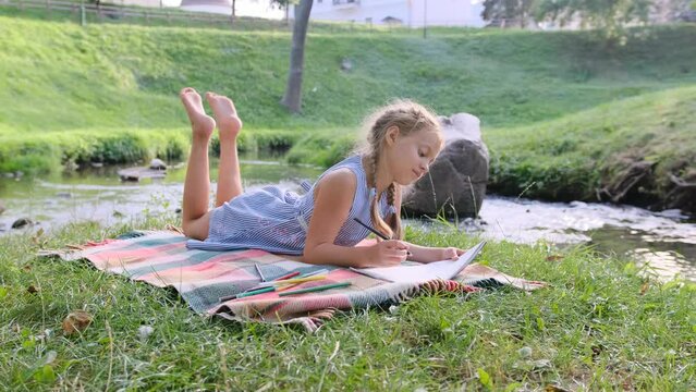 a young girl is lying on the bank of a river drawing with pencils in an album. The child enjoys the sunny weather