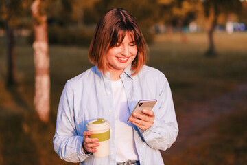 Photo of dreamy cute young lady wear blue shirt smiling walking enjoying tasty tea, coffee bamboo eco cup in green park and texting message  phone 5g