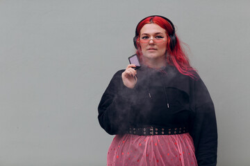 European plus size woman in headphones smoking vape electronic cigarette. Young red pink haired...