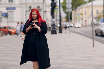 Fototapeta premium European plus size woman walking outdoor street city with mobile cellular phone. Young red pink haired body positive girl using cellphone smartphone