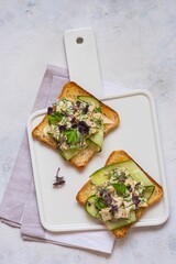 Open Danish smorrebrod sandwiches with chicken salad, fresh cucumber and herbs on a white ceramic board against a light concrete background. Sandwich recipes.