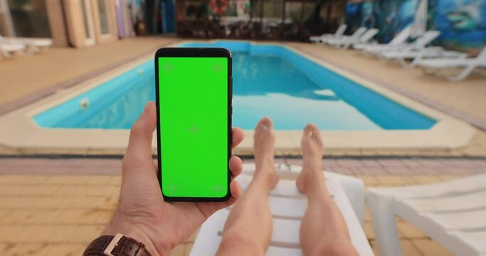 POV male hand holding smartphone with empty green screen chroma key relaxing near pool