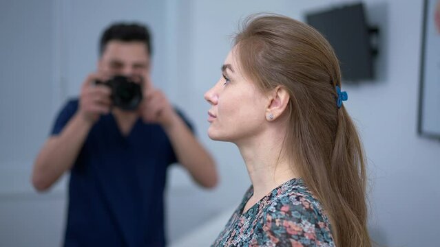 Side view slim adult Caucasian woman posing for before and after photo with blurred surgeon photographing with camera at background. Middle Eastern man taking photo of patient before plastic surgery