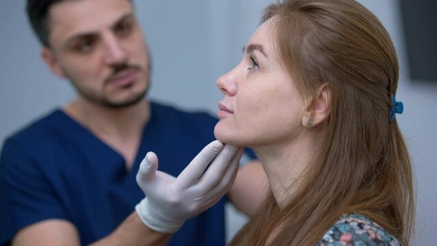 Side view face of beautiful Caucasian woman with blurred Middle Eastern plastic surgeon examining imperfections talking at background. Concentrated attractive lady consulting professional doctor