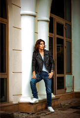 a young, handsome man with long dark hair, wearing a leather jacket, jeans and a white T-shirt, walks alone in the city