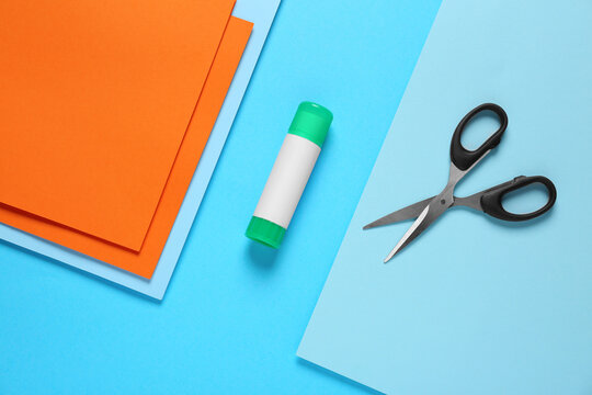 Glue stick, scissors and colorful paper on light blue background, flat lay
