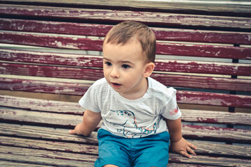 Bebé niño expresivo sentado en un banco de madera - obrazy, fototapety, plakaty