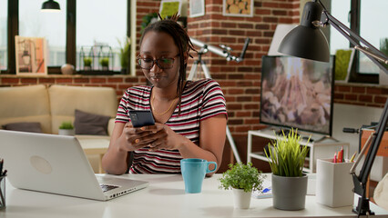 Company employee using smartphone to text messages on social media app, taking break from remote work at laptop. Young woman texting on mobile phone, working remotely at home desk.