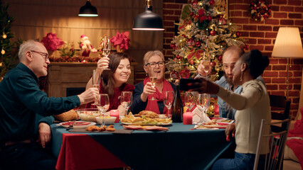 Joyful multiracial family members taking selfie photos with smartphone device while sitting at Christmas dinner table. Festive diverse people celebrating winter holiday together at home.