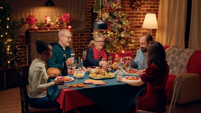 Laughing diverse people gathered around Christmas dinner table clinking wine glasses. Happy smiling heartily family members celebrating traditional winter holiday at home.