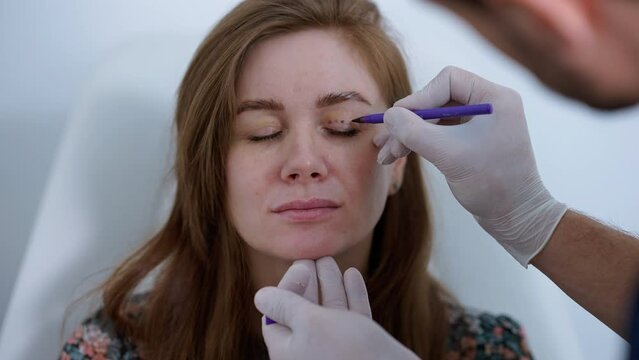 Caucasian woman with closed eyes in medical clinic with Middle Eastern plastic surgeon making eyelids for skin tightening. Unrecognizable concentrated doctor consulting patient