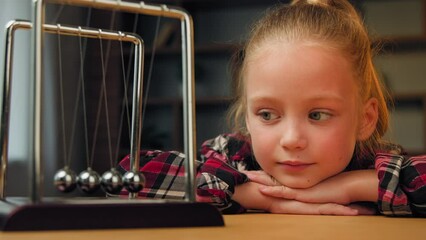 Concentrated child girl female little patient blonde cute beautiful small lady preschooler sitting on psychotherapy session indoors looking at swinging metal steel balls law kinetic energy hypnotized