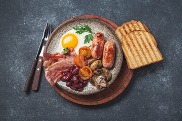 Traditional full English breakfast with fried eggs, sausages, beans, mushrooms, grilled tomatoes, bacon and toasts on gray plates.