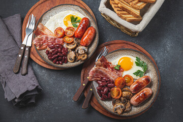 Traditional full English breakfast with fried eggs, sausages, beans, mushrooms, grilled tomatoes, bacon and toasts on gray plates.