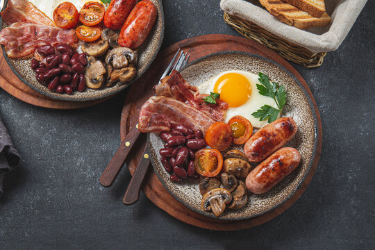 Traditional full English breakfast with fried eggs, sausages, beans, mushrooms, grilled tomatoes, bacon and toasts on gray plates.