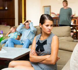 Upset young woman sitting at sofa during quarrelling between family on background at home