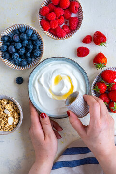 Woman Hands Pouring Maple Syrup Over Greek Yogurt, Raspberries, Strawberries, Blueberries And Granola On The Side.