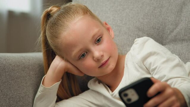Relaxed blonde child little girl blogger cute kid teen resting on couch playing online games on mobile phone posting photos in social media apps chatting with friends watching funny cartoons at home