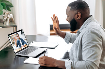 Online video meeting. African American young successful man talking on video conference with his colleague, friend or mentor while sitting at the desk in the office, talking about strategy,takes notes - obrazy, fototapety, plakaty