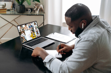 Fototapeta na wymiar Online training, lecture, seminar. African american smart student listening to the online lecture, taking notes in notebook, on the laptop screen are teacher and the group of multiracial students