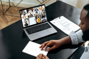 African american male smart student listening to the online lecture, taking notes in notebook, on...