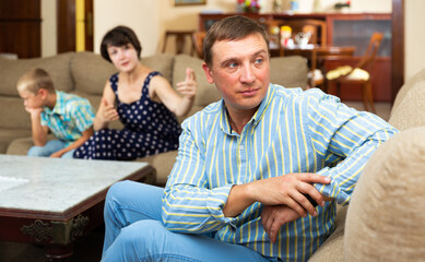 Unhappy man sitting at sofa after family quarrelling, woman with son on background