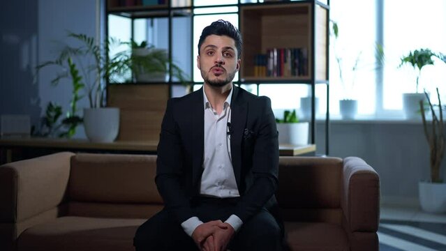 Portrait of serious confident businessman in elegant suit talking looking at camera sitting in modern office. Handsome Middle Eastern man posing blogging indoors. Talking head concept