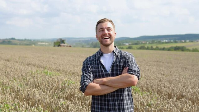 Happy young farmer standing on wheat field while combine harvester in background