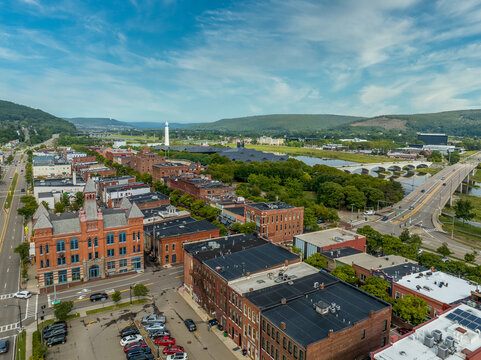 Aerial view of Corning Steuben County, New York downtown, Market Street, glass factory, chemung river, centerway walking bridge, little joe tower, parking lot with cloudy blue sky