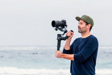 mature male on the beach holding a camera on a gimbal or stabilizer filming - obrazy, fototapety, plakaty