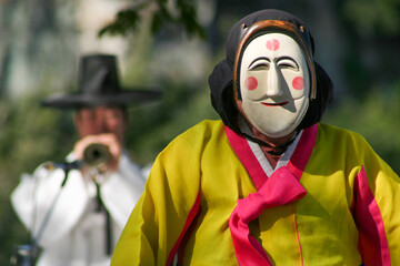 Person wearing a traditional South Korean mask while another man plays a tradtional instrument in the background