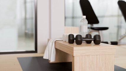 Fitness gym background with dumbbells, bottle of water, towel on a wooden seating bench.